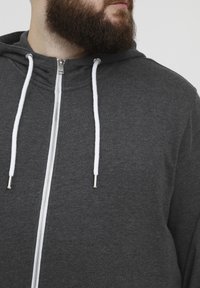 Sudadera con cremallera gris hecha de un material suave, con cordones blancos con puntas metálicas y una superficie texturizada.