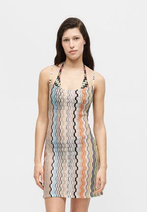 Kobieta w dopasowanej, bezrękawnikowej sukience z pionowymi falistymi paskami w kolorach niebieskim, pomarańczowym, czarnym i beżowym, stojąca na białym tle.