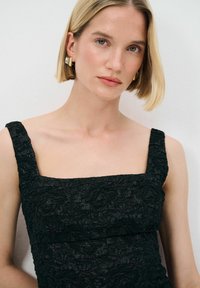 Schwarzes, strukturiertes Kleid mit quadratischem Ausschnitt und breiten Trägern, das ein erhabenes Blumenmuster aufweist. Einfaches Design ohne sichtbare Verschlüsse.