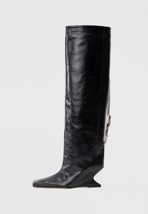 Botte noire en cuir jusqu'au genou avec un bout pointu, un talon compensé unique et un accent de chaîne latérale. Texture lisse avec une finition brillante.