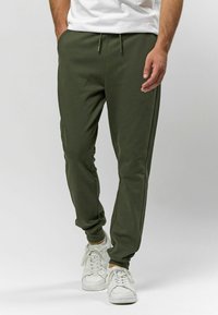 Olivové nohavice vyrobené z mäkkého materiálu, s elastickým pásom a šnúrkou, bočnými vreckami a prispôsobenými manžetami na členkoch.