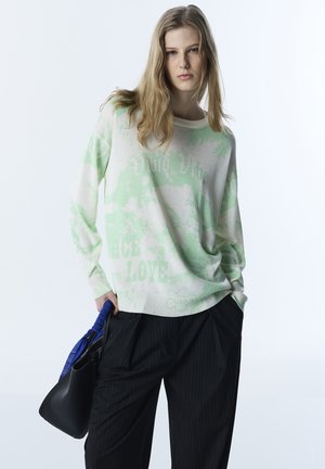 Młoda kobieta o długich blond włosach, ubrana w luźny, jasnooliwkowy i biały sweter oraz czarne spodnie w prążki, trzymająca czarną torebkę z niebieskim uchwytem.