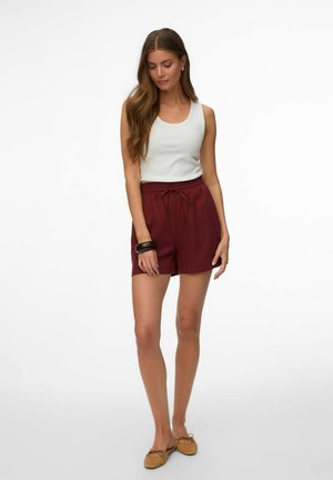 Femme portant un haut blanc sans manches, un short bordeaux à cordon, des chaussures plates couleur beige et un bracelet marron foncé, debout devant un fond blanc.