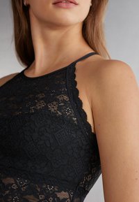 Černý krajkový top s vlnitými okraji, který se vyznačuje složitými květinovými vzory a tenkými ramínky. Hladký povrch zvýrazňuje elegantní design.