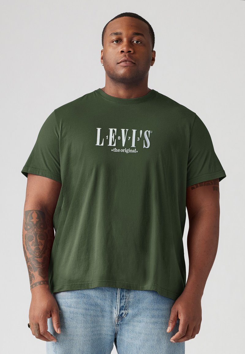 Alyvuogių žalia medvilnės marškinėliai su baltu Levi's logotipu. Trumpomis rankovėmis ir apvaliu kaklu, dėvima su šviesiai mėlynomis džinsais.
