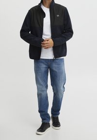 Man draagt een zwart en marineblauw jack met rits over een wit shirt, blauwe spijkerbroek en zwarte sneakers met witte zolen, staat met gevouwen handen.