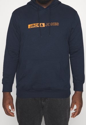 Mężczyzna ubrany w granatową bluzę z kapturem z pomarańczowym logo "JACK & JONES", w połączeniu z ciemnoszarymi spodniami, stoi na białym tle.