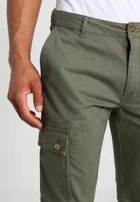 Olijfgroene cargobroek van katoen met een voorzak, zichtbare stiksels en een knoopsluiting bij de taille. Textuurstof.