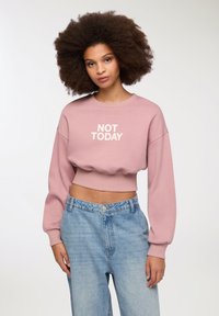 Felpa cropped rosa con il testo "NOT TODAY" in bianco. Presenta maniche lunghe e voluminose e una vita a coste. Abbinata a jeans azzurri.