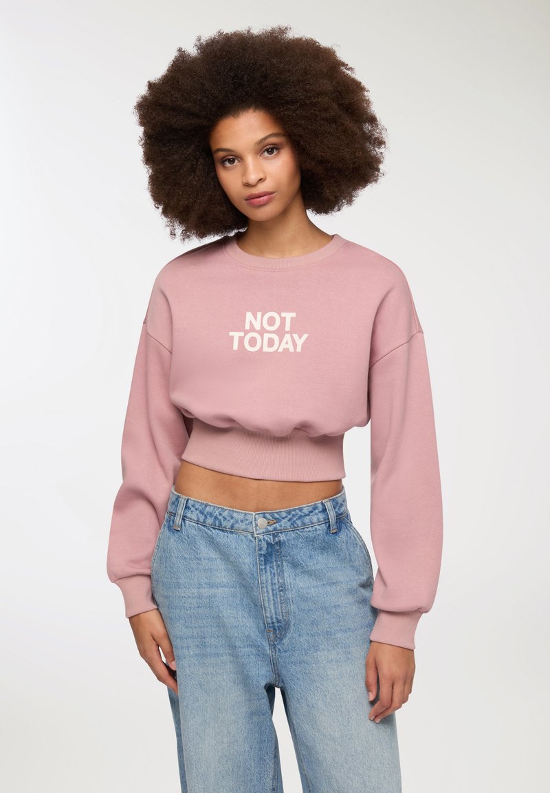 Felpa cropped rosa con il testo "NOT TODAY" in bianco. Presenta maniche lunghe e voluminose e una vita a coste. Abbinata a jeans azzurri.