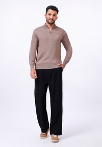 Beige pullover dengan kerah resleting dan logo kecil berwarna kuning, dipadukan dengan celana hitam longgar dan sepatu sneaker coklat muda. Tekstur yang halus.