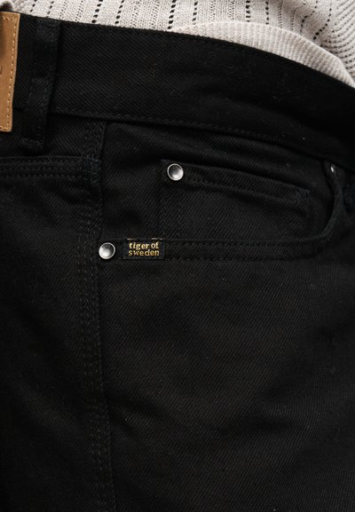 Κοντινό πλάνο τσέπης από μαύρο τζιν με μεταλλικά καρφιά και μια μικρή μαύρη και χρυσή ετικέτα "tiger of sweden".