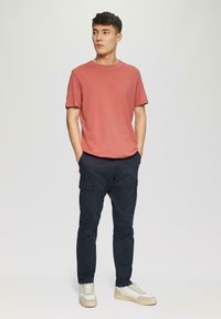 Rode t-shirt met ronde hals, marineblauwe cargobroek met zijzakken en witte sneakers met grijze accenten, staande op een lichte achtergrond.