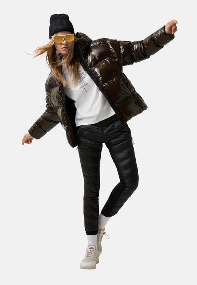 Kvinde iført sort hue, store gule solbriller, lysebrun skinnende pufferjakke, sorte quiltede bukser, hvide sokker og beige sneakers i en dynamisk pose.