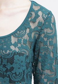 Teal spetsblus med ett blommönster, genomskinlig textur och rund halsringning med en slät kant. Designen har långa ärmar och intrikata detaljer.