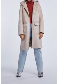 Beige chodící srstěný kabát s kapucí přes rezavý rolák, světle modré roztrhané džíny a černobílé vzorované kotníkové boty na stojící osobě.