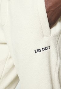 Casaco de fleece de cor creme com o logótipo bordado "LES DEUX", apresentando uma superfície texturizada e um detalhe em couro marrom liso na manga.