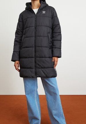 Personne portant un manteau matelassé à capuche noir Adidas avec une fermeture éclair à l'avant et un jean bleu clair, debout sur un sol brun.