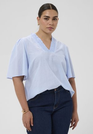 Jeune femme aux cheveux foncés portant un chemisier bleu clair à manches courtes et un jean foncé, sur un fond gris uni.