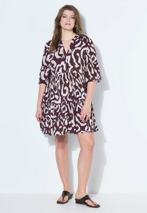 Frau trägt ein lockeres, knielanges Kleid mit braun-weißem abstraktem Muster, Dreiviertelärmeln und schwarzen Keilsandalen.