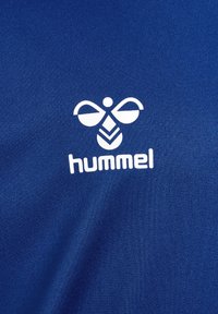 Tejido atlético azul con un logo blanco que se asemeja a una abeja, con alas simétricas y dos formas de hoja apuntando hacia abajo en el centro.