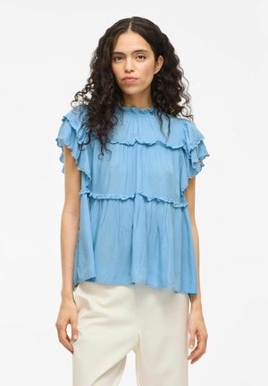 Himlenblå blus med lager av ruffle-detaljer, korta ärmar och rund halsringning, tillverkad av ett lätt material.