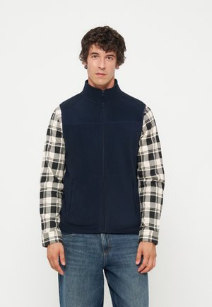 Homme portant un gilet zippé bleu marine par-dessus une chemise à carreaux noir et blanc avec un jean bleu, debout devant un fond clair uni.