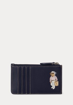 Suport pentru carduri din piele albastru marin cu cusături albe și un urs mic care poartă ochelari de soare, o cămașă cu dungi, pantaloni albi și poartă o geantă bej.