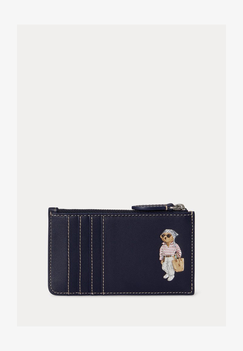 Porte-cartes en cuir bleu marine avec des coutures blanches et un petit ours portant des lunettes de soleil, une chemise rayée, un pantalon blanc et un sac beige.