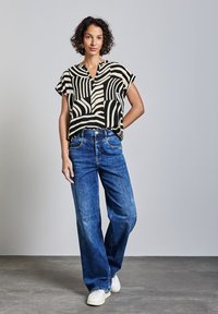 Blusa a maniche corte a righe nere e crema con scollatura a V, abbinata a jeans in denim blu larghi e sneakers bianche su uno sfondo grigio.