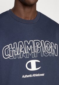 Βαμβακερό μπλουζάκι σε ναυτικό μπλε χρώμα με έντονη λευκή επιγραφή «CHAMPION» και λογότυπο. Λείο ύφασμα, ριμπ λαιμόκοψη και λεπτομέρεια «Authentic Athleticwear».
