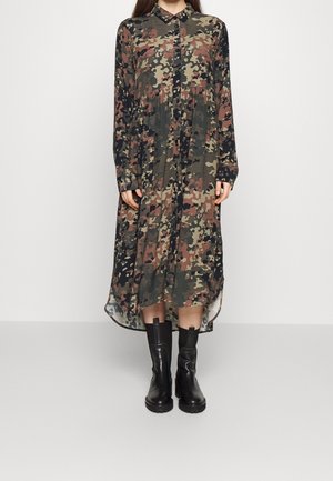 Langes Hemdkleid mit Camouflage-Muster in verschiedenen Grüntönen, Braun und Schwarz. Mit klassischem Kragen und langen Ärmeln. Passt gut zu schwarzen Stiefeln.
