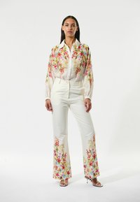 Floral-patterned, sheer blouse in white with pink and green embroidery, paired with fitted white pants featuring a similar floral design at the hems.