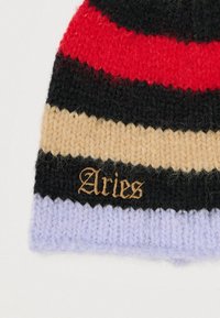 Ριγέ πλεκτό ύφασμα σε κόκκινο, μαύρο, μπεζ και λεβάντα, με το "Aries" κεντημένο σε χρυσά γοτθικά γράμματα κοντά στην κάτω άκρη.