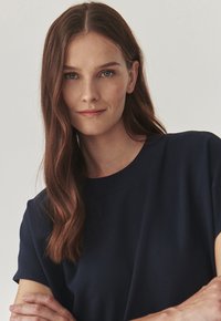 Haut à manches courtes bleu marine avec un col rond, fabriqué dans un tissu doux. Le modèle a les cheveux bruns longs et ondulés et les bras croisés.