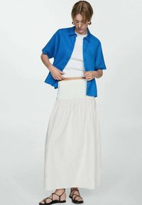 Camicia blu a maniche corte sopra un top corto bianco, abbinata a una lunga gonna bianca arricciata. Sandali neri con cinturino completano l'outfit.