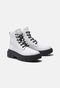 Bottines en cuir blanc avec lacets noirs, semelle noire épaisse et coutures renforcées, arborant un logo sur le côté extérieur.