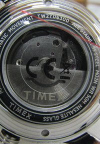 Caja de reloj de acero inoxidable con un fondo transparente que muestra un mecanismo de movimiento. El texto impreso incluye "TIMEX" y la certificación CE.