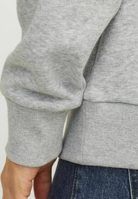 Grijze sweatshirt met een zachte textuur, geribbelde mouwen en onderkant. Zichtbare hand die de stof bij de pols vasthoudt, in combinatie met donkere denim.