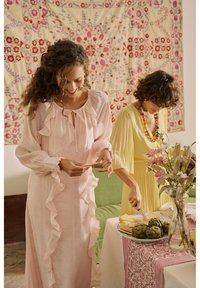Deux femmes en robes pastel préparent de la nourriture à une table avec des artichauts, du fromage et des fleurs, debout dans une pièce décorée.