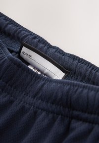 Temno modri elastični hlačniki z všito belo etiketo z napisom "NAME" in prazno vrstico za pisanje.