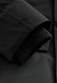 Manga de chaqueta negra que presenta una tela suave y mate con un puño de punto negro acanalado. El diseño es sencillo, sin patrones o acentos visibles.