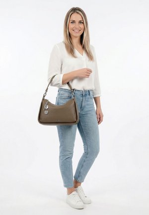 Woman in white blouse and blue jeans holds taupe leather handbag with silver charms, smiling against white background.