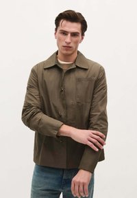 Lichte bruine jas met een kraag, voor knopen en een borstzak. Draagt over een beige gebreide trui en blauwe spijkerbroek.