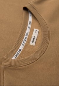Sweat-shirt marron en coton à col rond côtelé avec des étiquettes de marque blanches visibles indiquant "ombre cotton line" et taille "M".