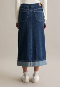 Długa spódnica jeansowa w ciemnym niebieskim kolorze z podwiniętym dołem, wyposażona w boczne kieszenie i metkę na tylnej części pasa, zestawiona z białymi trampkami i jasnymi skarpetkami.