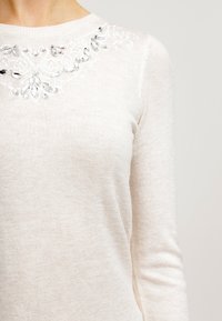 Pull blanc à manches longues, présentant un tricot texturé et des embellissements décoratifs en perles autour de l'encolure.