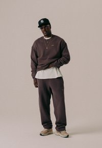 Dunkelbrauner, oversized Sweatshirt mit Rundhalsausschnitt, passende Jogginghosen und helles Schuhwerk. Zubehör umfasst eine schwarze Kappe und mehrschichtige Halsketten.
