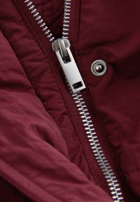 Gros plan d'une fermeture éclair en métal argenté partiellement fermée sur une veste en tissu bordeaux avec un bouton-pression métallique visible.