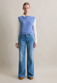 Jasnoniebieski sweter bez rękawów o teksturowanym wzorze na białej koszuli z długim rękawem, zestawiony z jasnoniebieskimi, szerokimi dżinsami o wysokim stanie oraz brązowym obuwiem.